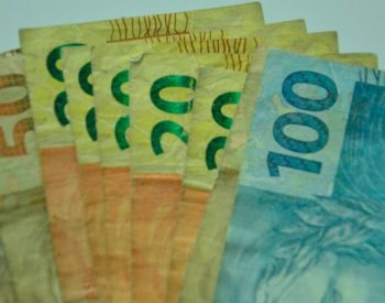 Cédulas de dinheiro brasileiro de diferentes valores estão sobrepostas e espalhadas sobre uma superfície clara.
