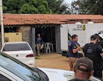 Agentes da Polícia Civil e da perícia estão reunidos diante de um imóvel, ao lado de viaturas e de um veículo da Polícia Científica